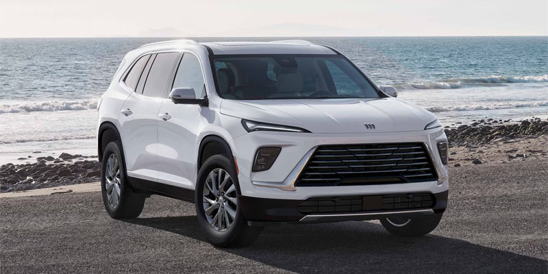 A white 2026 Buick Enclave sits on the stony beach with a low tide washing in from the soft blue waves